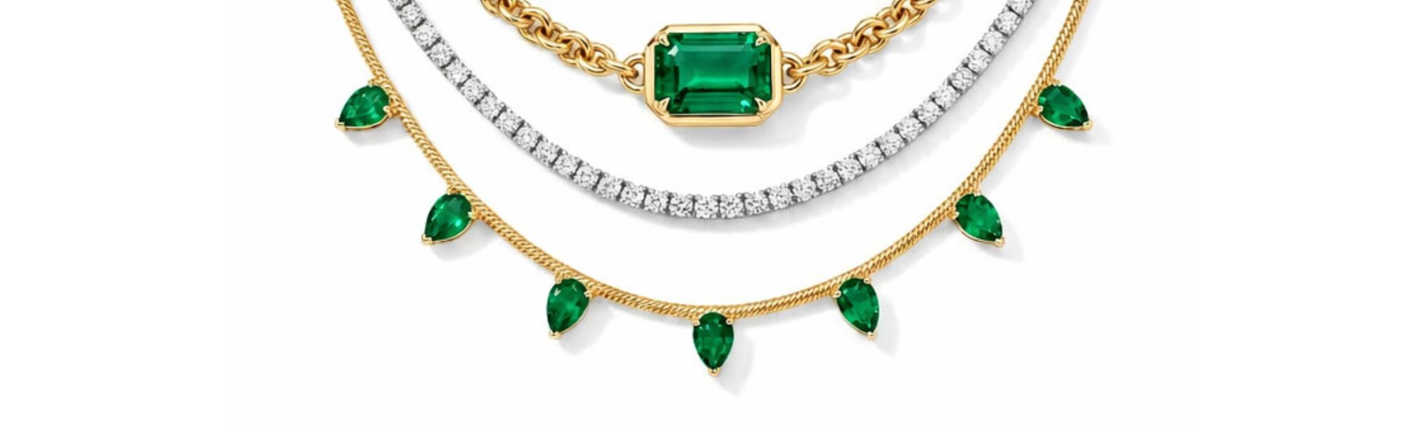 Three layered necklaces: a gold chain with a rectangular green gemstone, a silver tennis necklace with clear stones, and a gold necklace with seven dangling teardrop-shaped green gems.