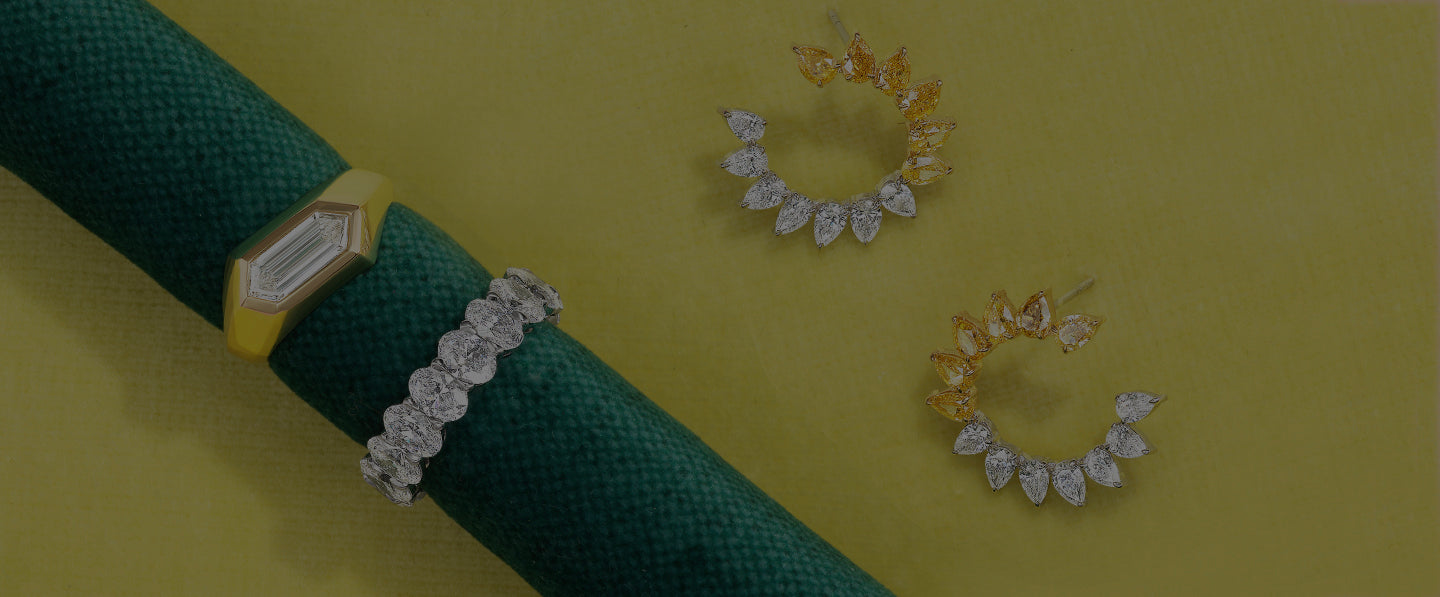 A gold ring with a geometric diamond, a silver bracelet with round diamonds on a green cloth, and two pairs of earrings with teardrop-shaped stones on a yellow background.