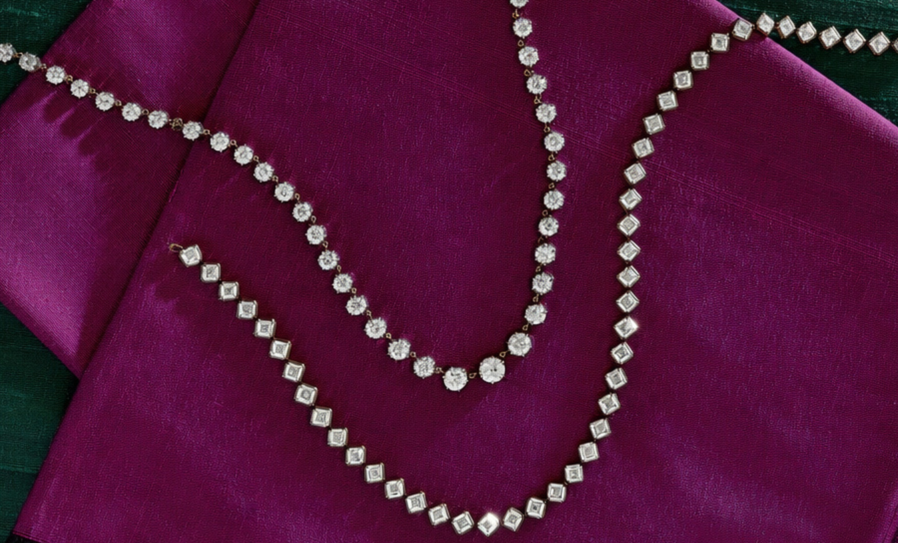 Two gold chain necklaces displayed on a rich burgundy fabric; one chain is thicker with a pink gemstone pendant, the other is beaded with a round clasp.