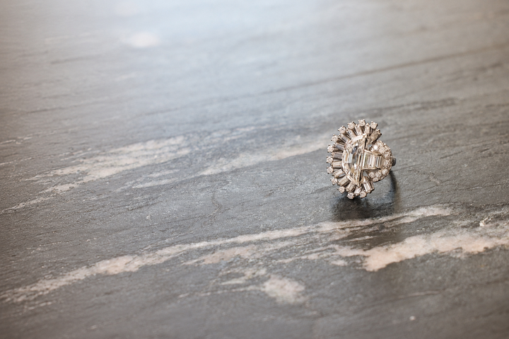 A diamond cluster ring with a circular design sits upright on a gray, marbled surface. The rings intricate setting sparkles in the light, while the background remains softly blurred.