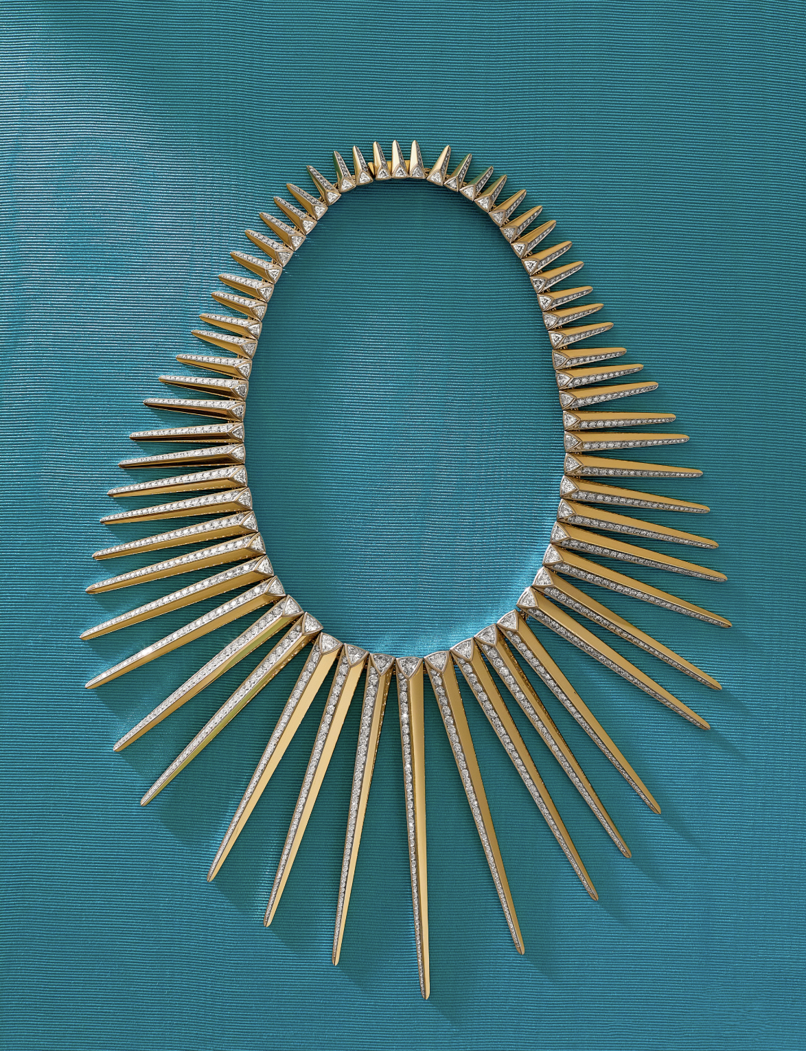 A gold necklace with long, pointed spikes radiating outward, some spikes encrusted with diamonds, displayed on a turquoise textured background.