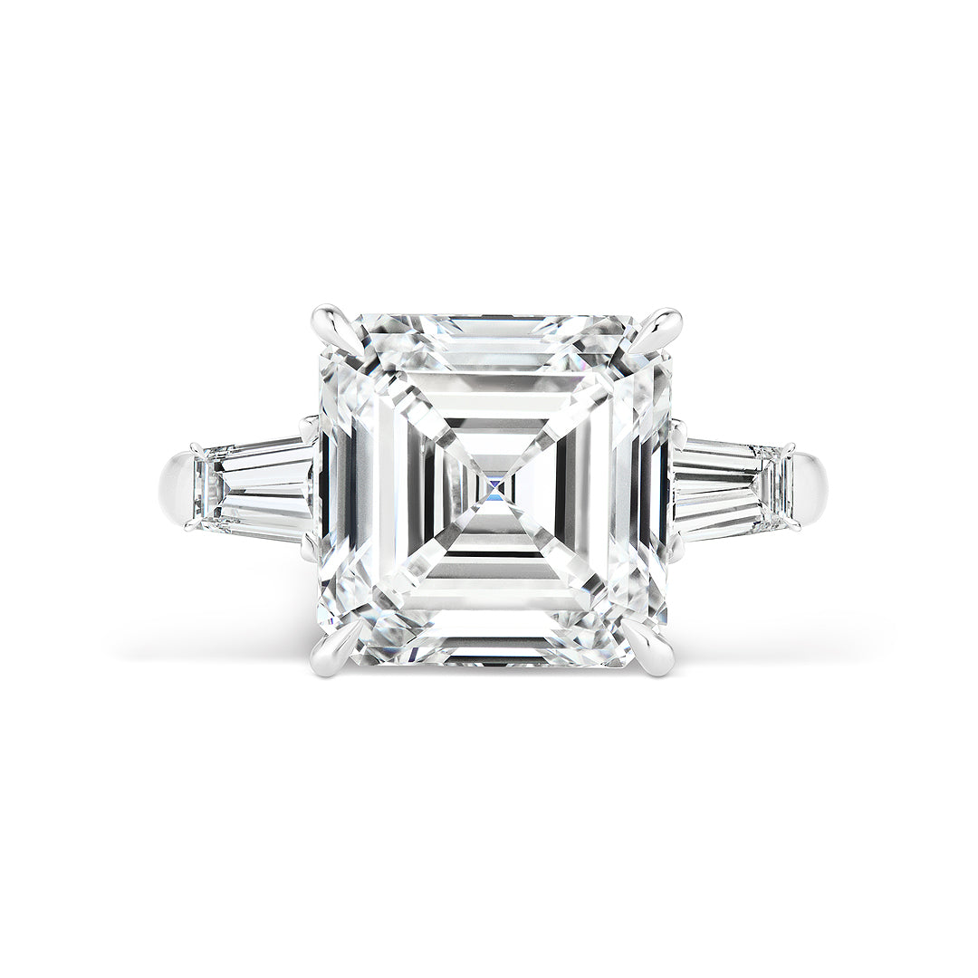 The Asscher Cut Diamond Engagement Ring features a platinum band with a central Asscher-cut diamond and two tapered baguette side stones, beautifully set against a white background.
