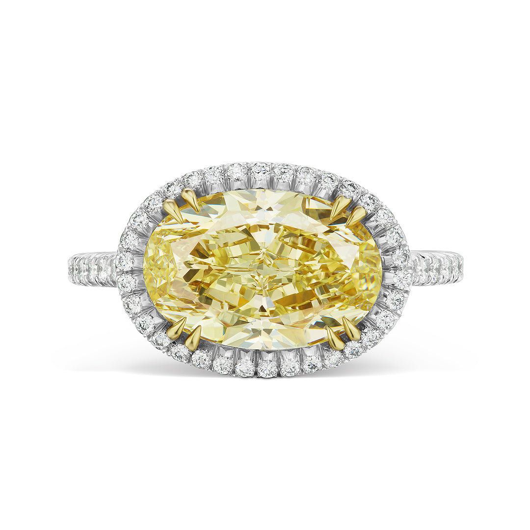 The East-West Fancy Yellow Oval Diamond Engagement Ring features a vibrant oval yellow diamond center, haloed by white diamonds, and set on a pavé-encrusted white metal band for a dazzling look.