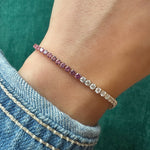 A wrist wearing the This & That Round Brilliant Diamond and Pink Sapphire Tennis Bracelet, featuring alternating gemstones in a slim 18k yellow gold band. The wearer is styled in a blue denim jacket with a green background.