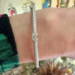 A close-up shows a wrist wearing the East-West Emerald Cut Diamond Pavé Bangle—an 18K white gold bangle with a central emerald cut diamond and pavé accents. The person sports a green and black checkered sleeve against a colorful, blurred background.