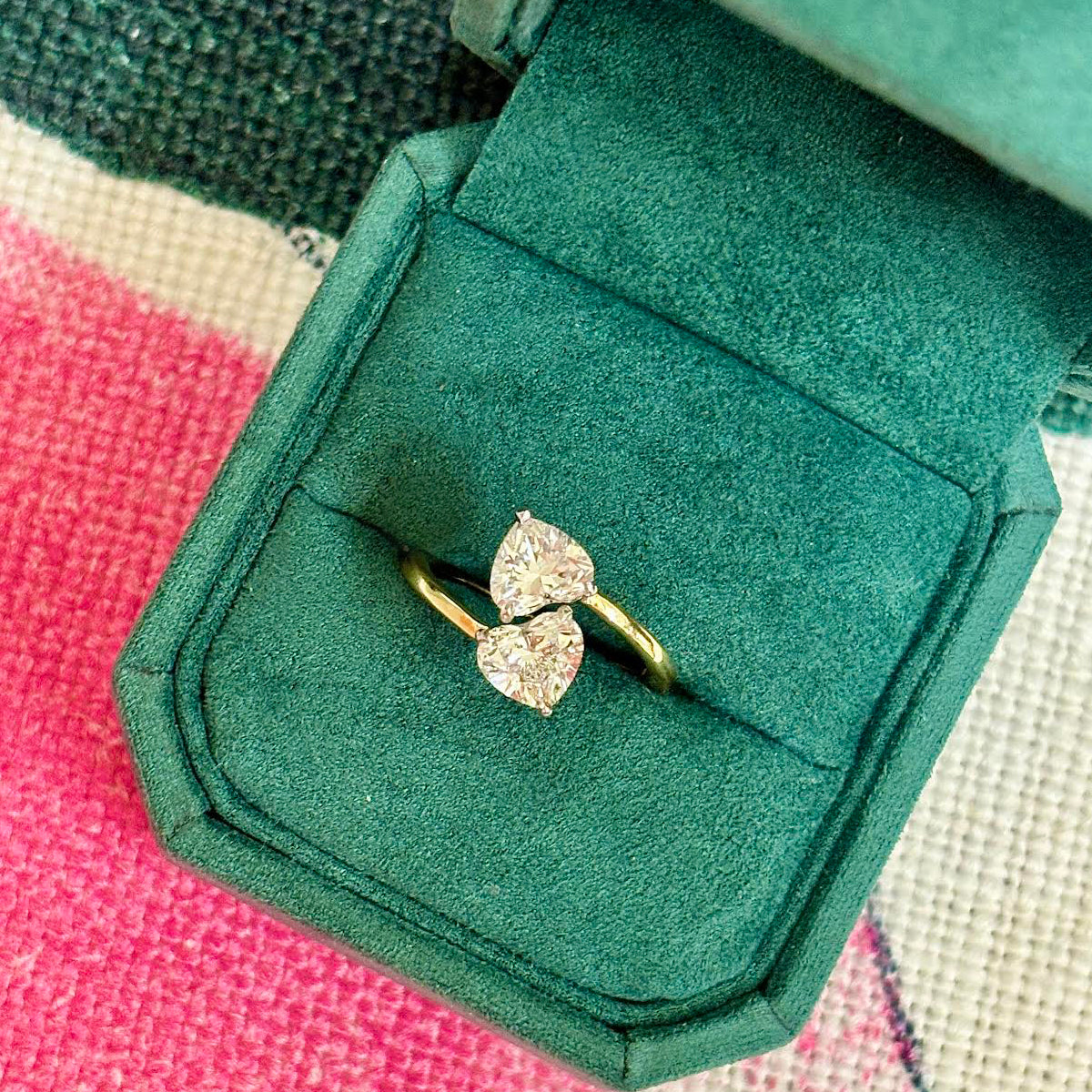 The Double Heart Wrap Around Ring, featuring 2.07tcw diamonds set in 18K yellow gold, is showcased in a green velvet box atop a vibrant, patterned surface.