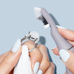 With pale blue nails, a person uses the Juli Diamond Cleaning Essentials Kit's brush and foam to clean a silver ring with a large gemstone against a light blue background.