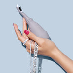 A hand with pink polished nails holds the Juli Diamond Cleaning Essentials Kit, wearing sparkling diamond rings and bracelets, all set against a light blue background.