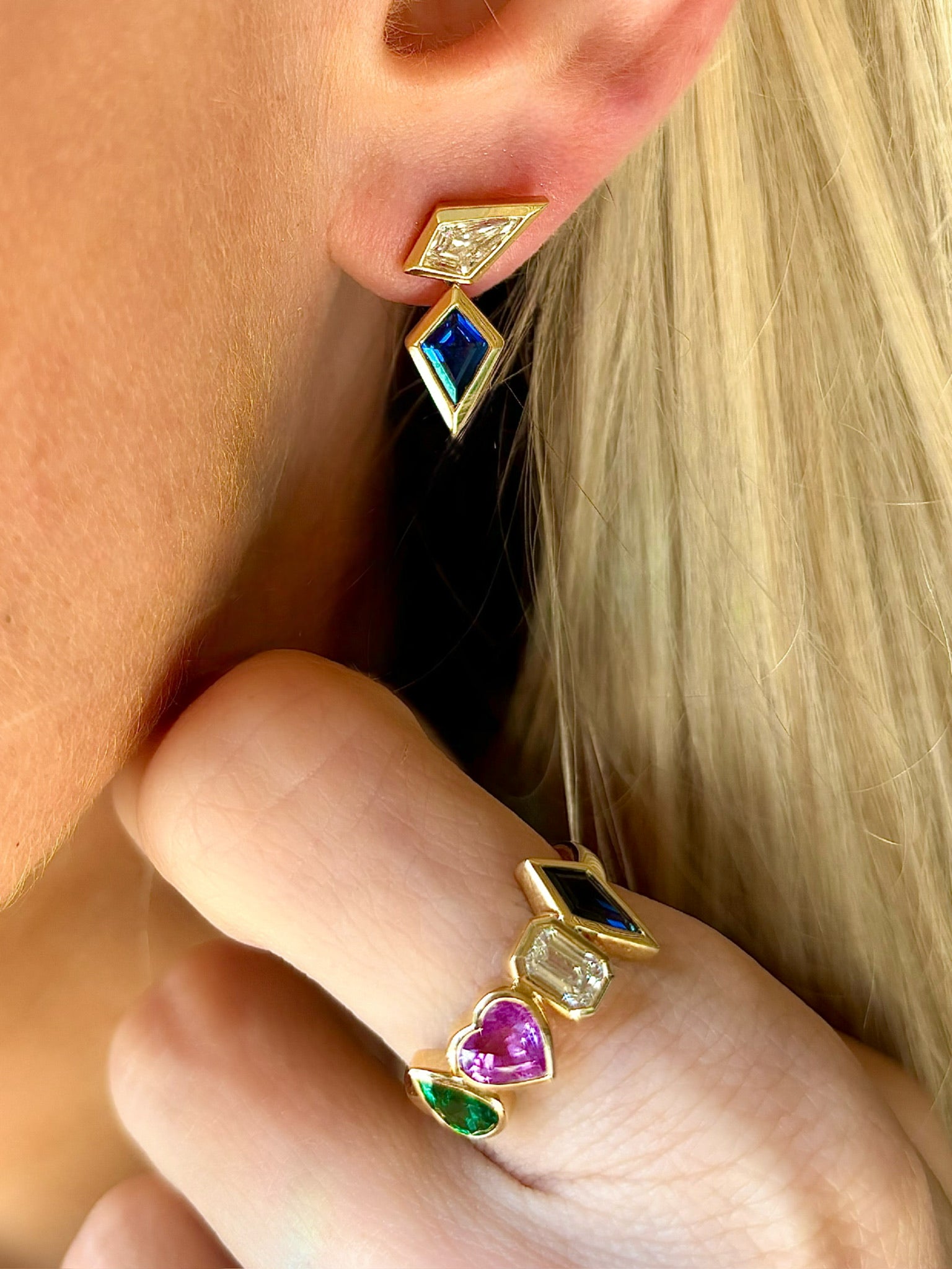 A close-up of a person wearing geometric gold earrings with blue and clear stones and the Chroma Heart Beats Bezel Set Cluster Ring in yellow gold, featuring emeralds, diamonds, and sapphires, with blonde hair in the background.