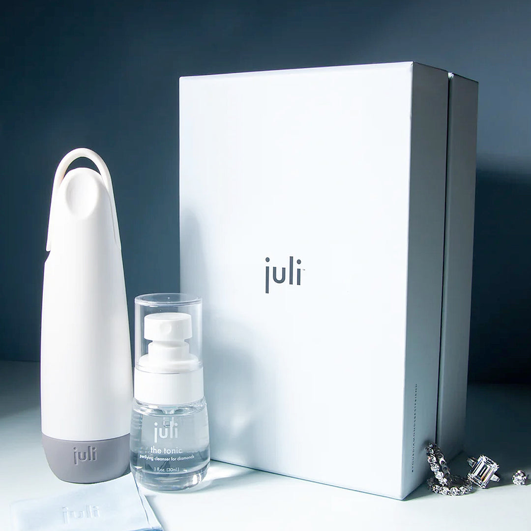 The Juli Diamond Cleaning Essentials Kit includes a diamond cleanser in a white handled bottle, a small foaming pump bottle, and a large white box, all displayed on a smooth surface with a silver chain nearby.