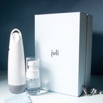 The Juli Diamond Cleaning Essentials Kit includes a diamond cleanser in a white handled bottle, a small foaming pump bottle, and a large white box, all displayed on a smooth surface with a silver chain nearby.