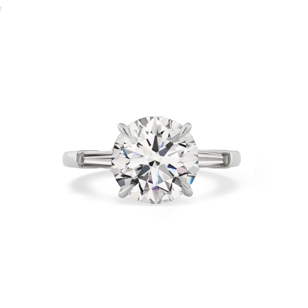 The Round Brilliant Diamond Engagement Ring with Tapered Baguette Side Stones features a central round diamond and two tapered baguette diamonds on each side, shown against a white background.
