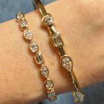 A close-up of a wrist wearing two East-West Bezel Set Mixed Shape Diamond Tennis Bracelets in yellow gold, each featuring various diamond shapes, set against a blurred neutral background.