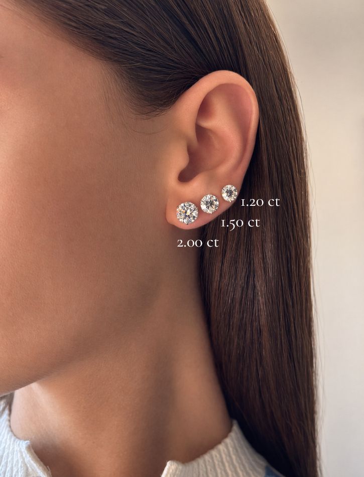 Close-up of a person’s ear adorned with three Classic Round Diamond Studs in Martini Setting, each featuring a different size—1.20 ct, 1.50 ct, and 2.00 ct—from smallest to largest on the earlobe. These stunning studs showcase GIA certified diamonds set in elegant 18k White Gold for a timeless look.