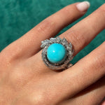 A hand displays the Turquoise and Diamond Ring: a platinum band featuring a large turquoise cabochon center, encircled by dazzling white stones, set against a teal background.
