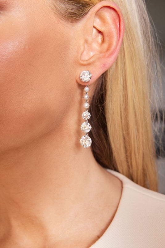 Close-up of a woman's ear wearing Bespoke Graduated Drop Ear Jackets in white gold, featuring pearls and round brilliant diamonds set in a vertical line. She has straight blonde hair and a beige top—an elegant customizable piece.