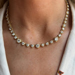 A close-up shows a person wearing the Collet Set Round Diamond Graduated Tennis Necklace in Yellow Gold, with sparkling prong-set diamonds around their neck. They are dressed in a white top with a small embroidered detail.