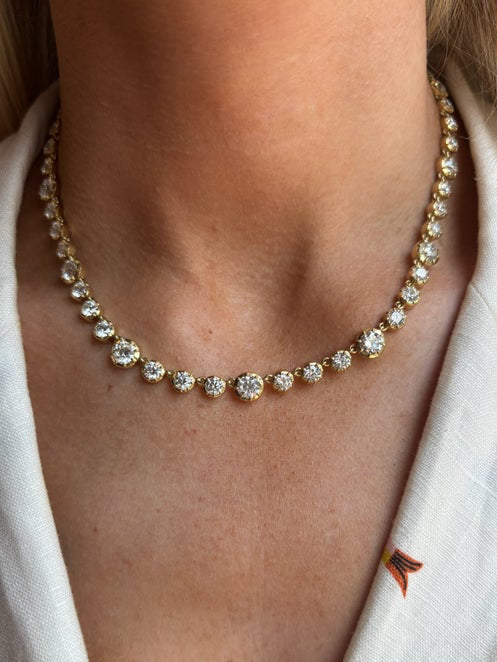 A close-up shows a person wearing the Collet Set Round Diamond Graduated Tennis Necklace in Yellow Gold, with sparkling prong-set diamonds around their neck. They are dressed in a white top with a small embroidered detail.