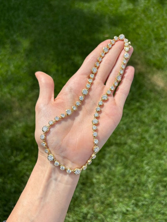 A hand holds the Collet Set Round Diamond Graduated Tennis Necklace in Yellow Gold, sparkling against a blurred green grass background—this elegant necklace shimmers beautifully in the sunlight.