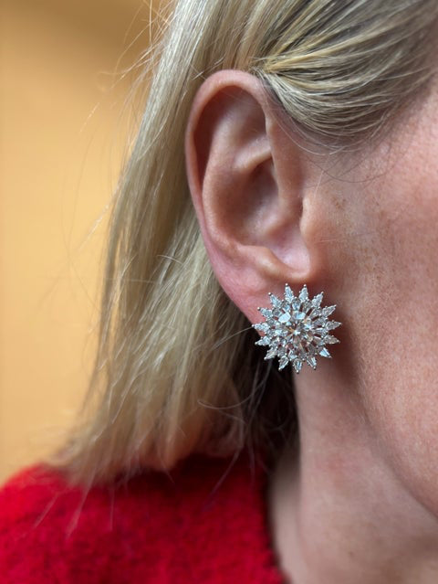 Close-up of a person with light hair in a red garment wearing Radial Shield Diamond Ear Jackets in White Gold, designed in a striking starburst shape, set against a warm background.
