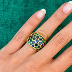 A hand wears the Sabbadini Emerald and Sapphire Ring in 18K yellow gold, set with sapphire and emerald gemstones, against a green textured background.