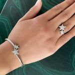 A hand rests on a green marble surface, wearing a silver bracelet and the Celestial Shooting Stars Wraparound Ring with Kite Diamonds, both adorned with sparkling star-shaped designs.