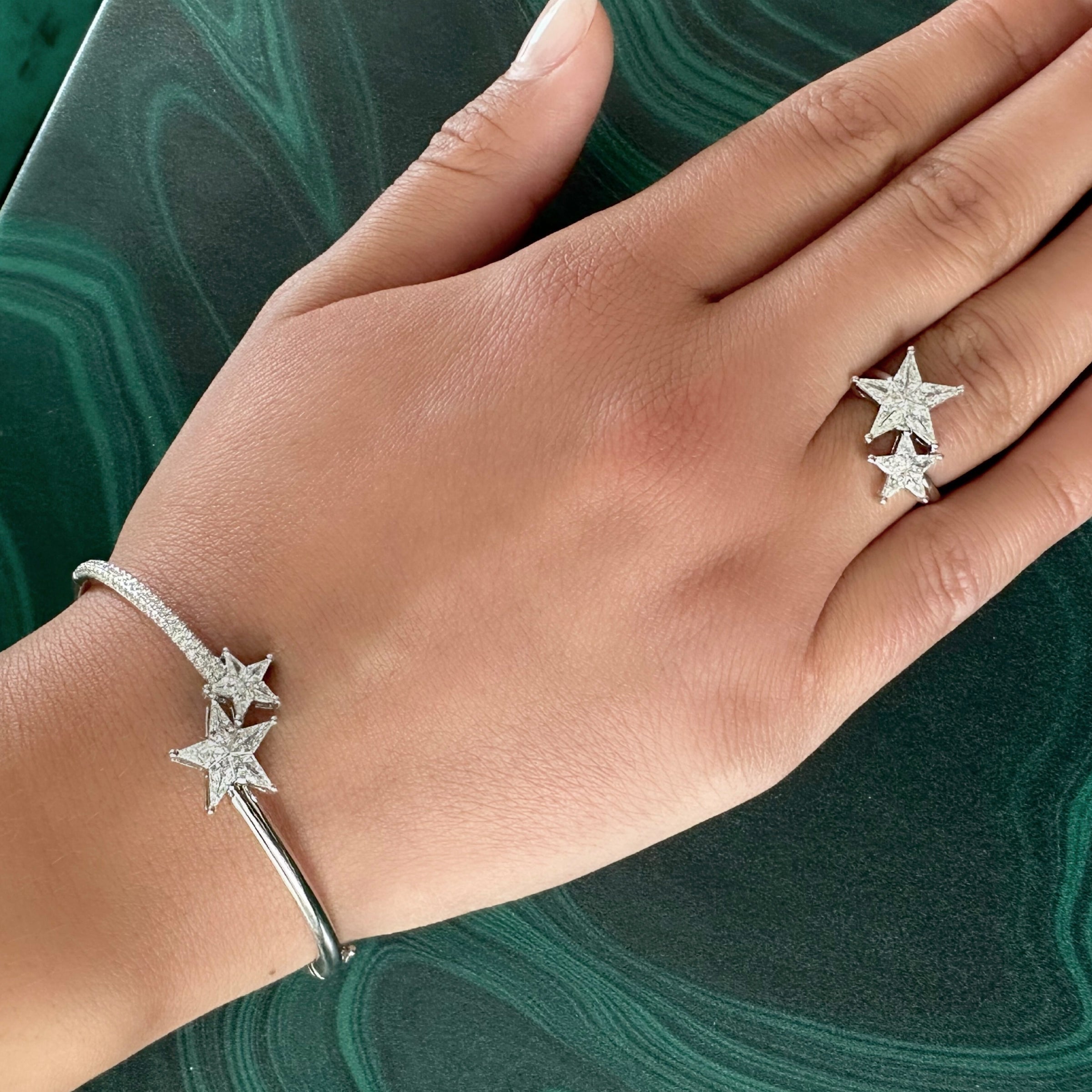 A hand rests on a green marble surface, wearing a silver bracelet and the Celestial Shooting Stars Wraparound Ring with Kite Diamonds, both adorned with sparkling star-shaped designs.