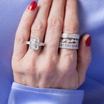 A hand with red-painted nails rests on light blue satin, wearing an Emerald Cut Diamond Eternity Band on the ring finger and three sparkling diamond celebration rings stacked on the middle finger.
