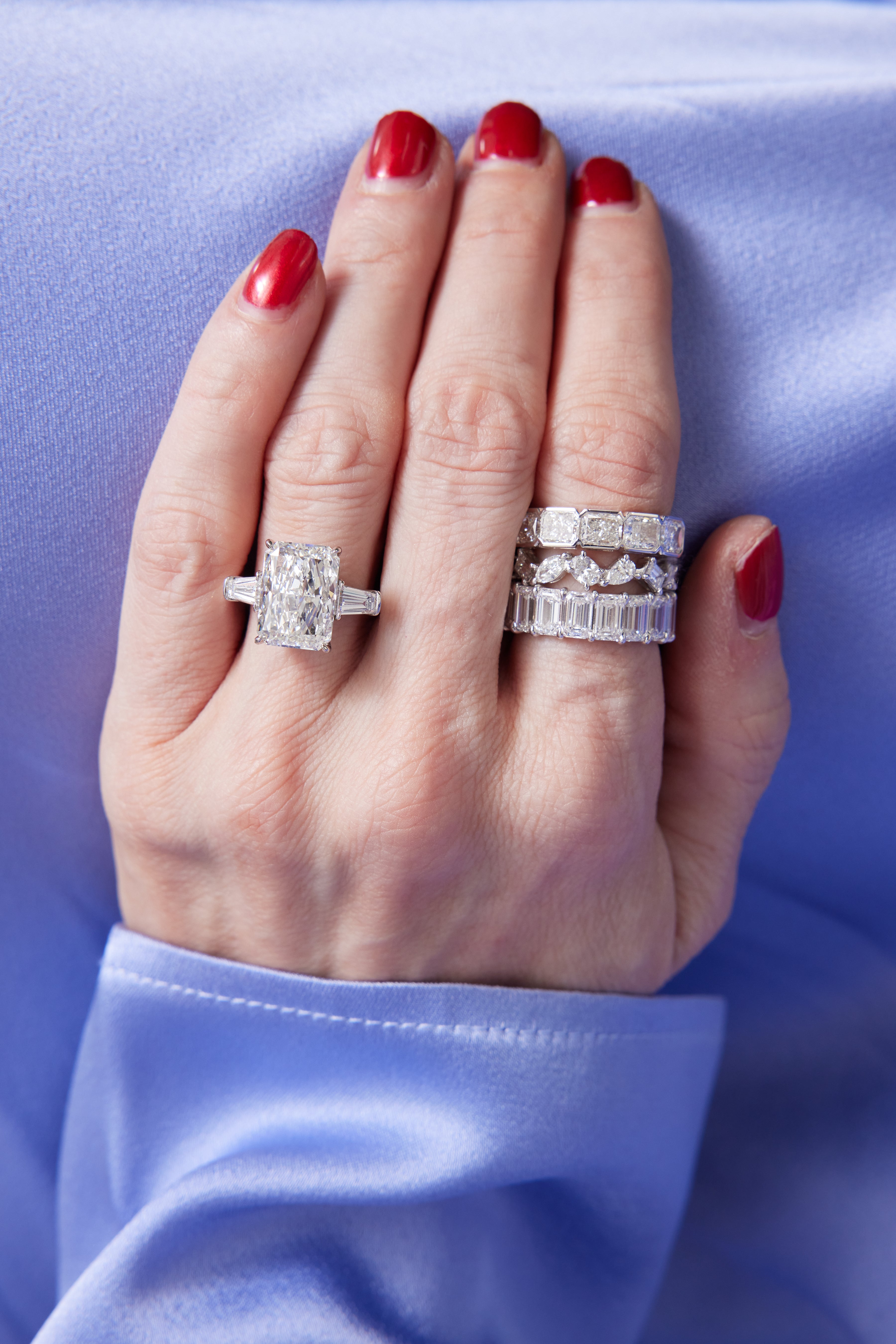 A hand with red-painted nails rests on light blue satin, wearing an Emerald Cut Diamond Eternity Band on the ring finger and three sparkling diamond celebration rings stacked on the middle finger.