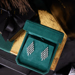 Art Deco Chandelier Earrings in white gold with emerald cut diamonds (VS1-VS2 clarity), shown in a green velvet box, surrounded by black and gold gift boxes and decorative accents.
