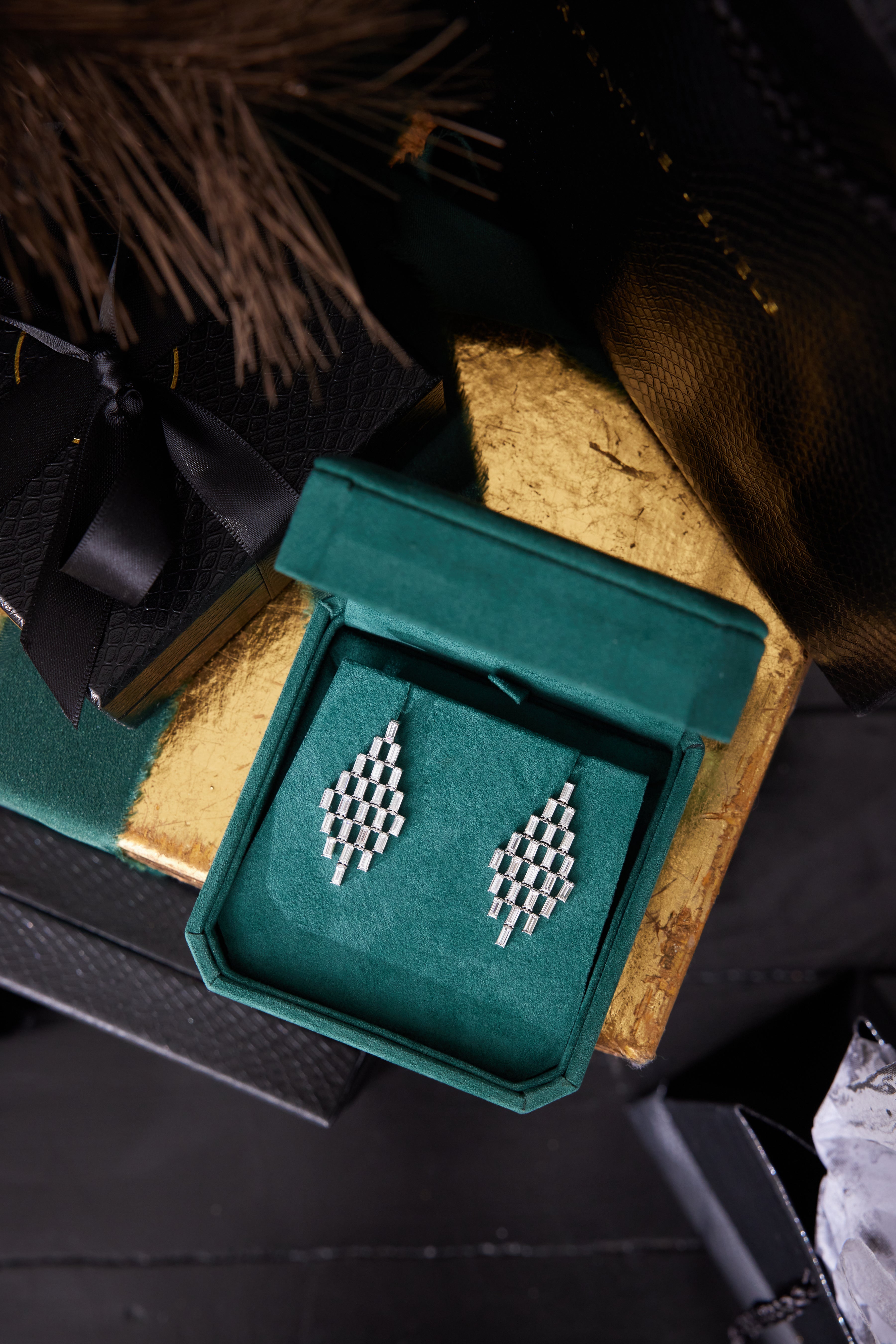 Art Deco Chandelier Earrings in white gold with emerald cut diamonds (VS1-VS2 clarity), shown in a green velvet box, surrounded by black and gold gift boxes and decorative accents.