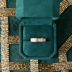 A Half Bezel Emerald Cut Diamond Eternity Band is displayed in an open green velvet ring box, set on a green and gold beaded fabric background.