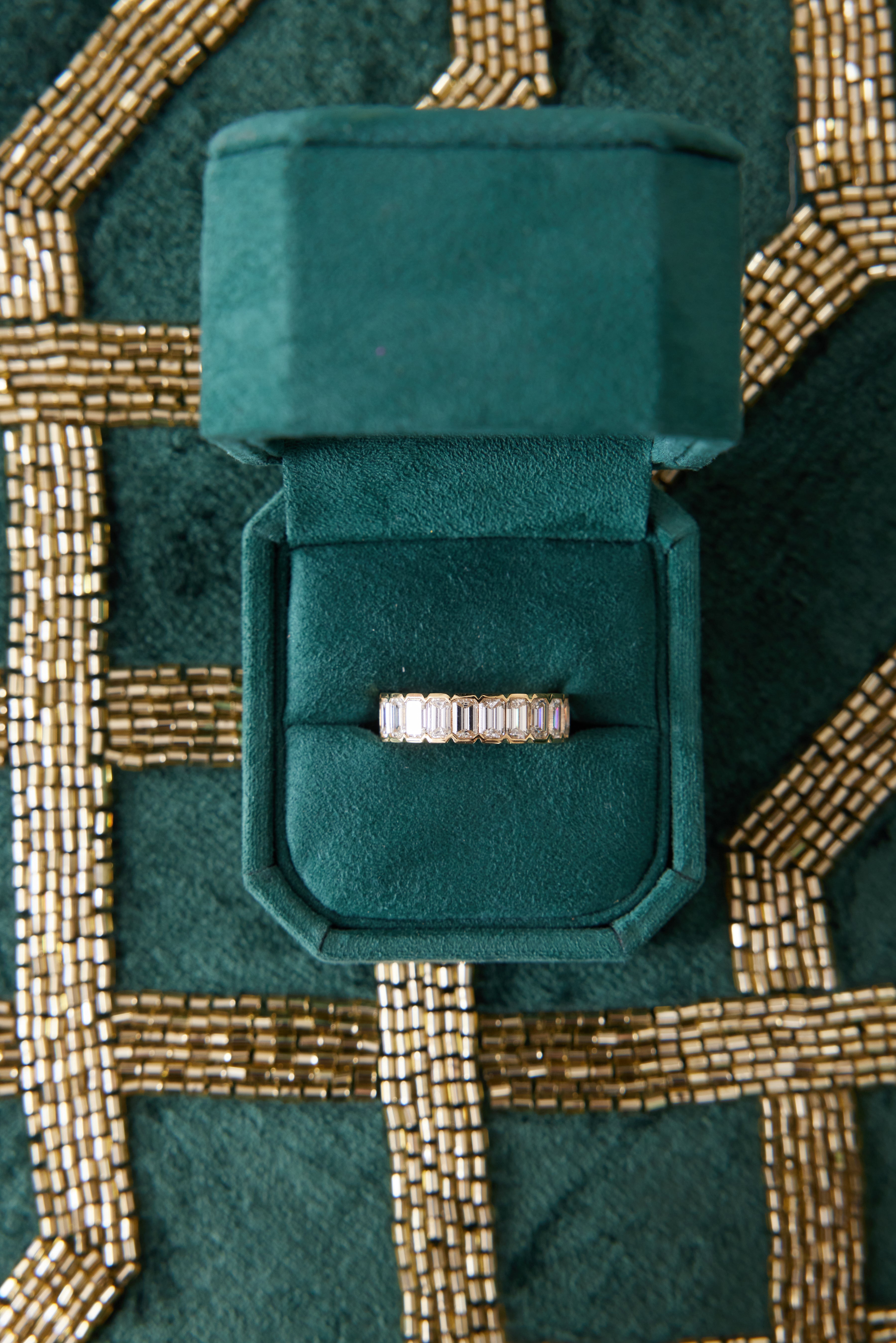 A Half Bezel Emerald Cut Diamond Eternity Band is displayed in an open green velvet ring box, set on a green and gold beaded fabric background.