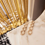 Chrysler Graduated Hexagon Drop Earrings in Yellow Gold with Baguette Diamonds, featuring interlocking hexagons, rest on a white marble surface with a blurred background and decorative golden chains.
