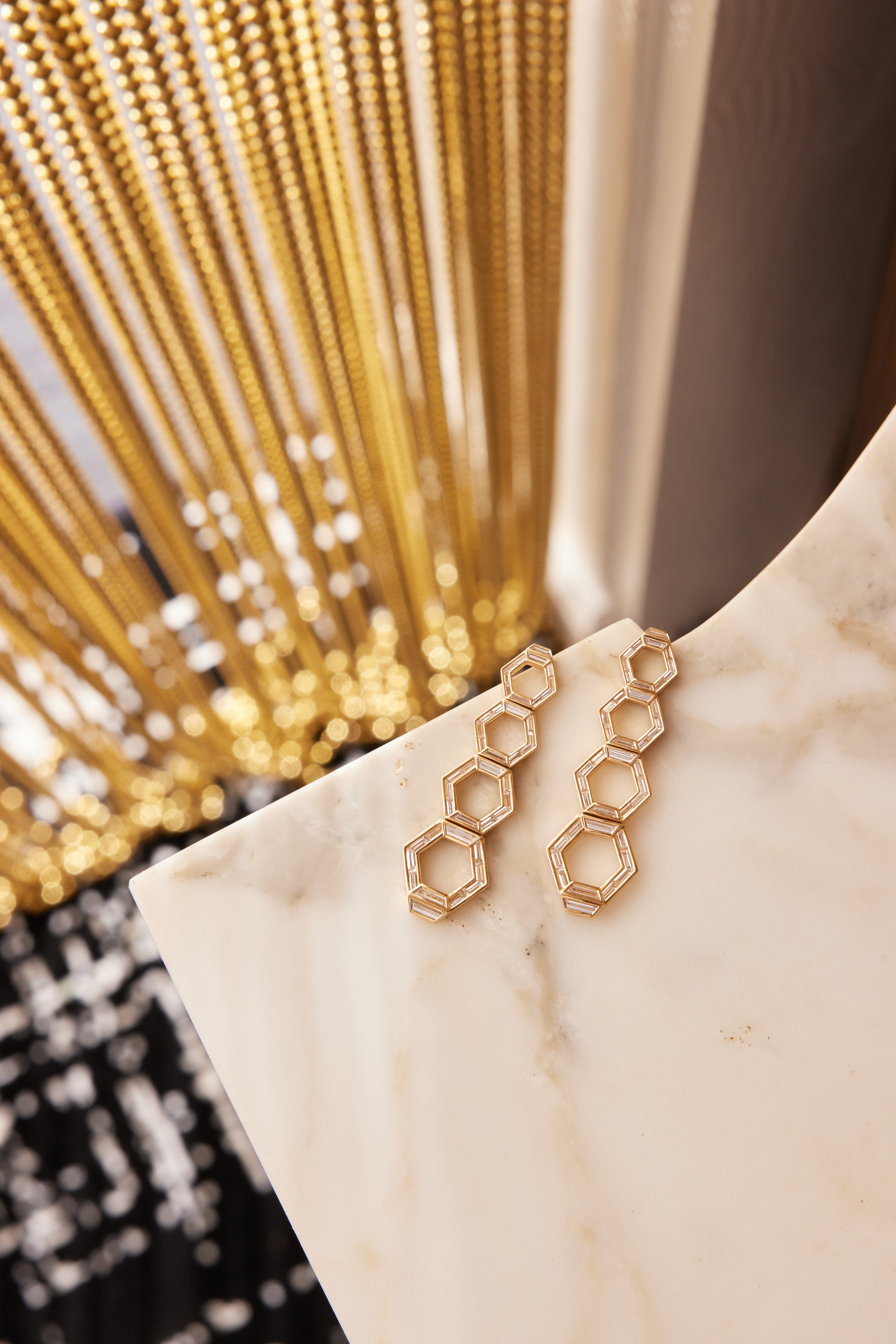 Chrysler Graduated Hexagon Drop Earrings in Yellow Gold with Baguette Diamonds, featuring interlocking hexagons, rest on a white marble surface with a blurred background and decorative golden chains.