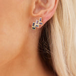 Close-up of a light-skinned person with blonde hair wearing Chevron Drop Earrings in yellow gold, featuring multiple vibrant multicolor baguette cut sapphires arranged in a geometric design.