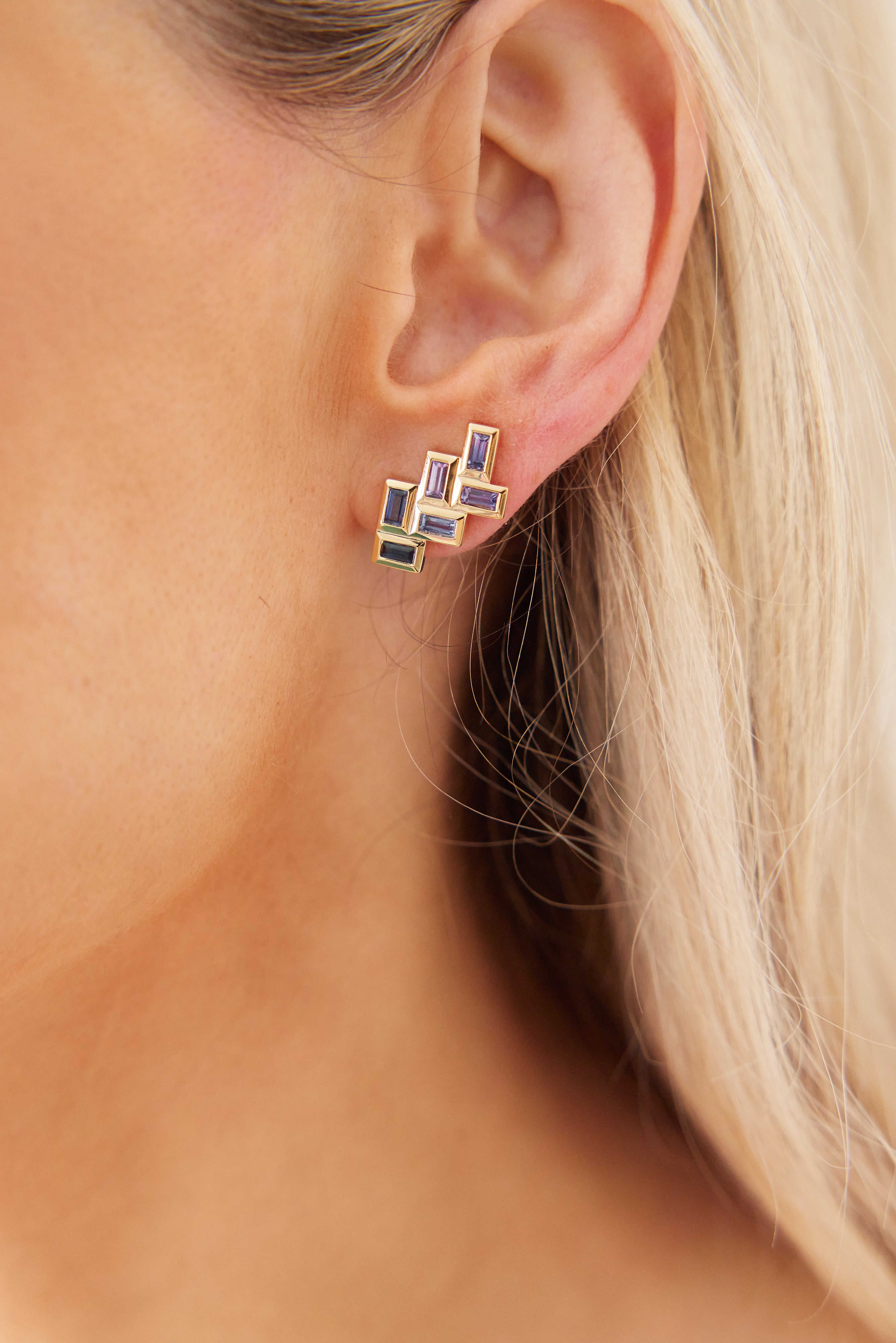 Close-up of a light-skinned person with blonde hair wearing Chevron Drop Earrings in yellow gold, featuring multiple vibrant multicolor baguette cut sapphires arranged in a geometric design.