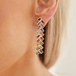 Close-up of a woman's ear wearing Chevron Drop Earrings in yellow gold, featuring a geometric zigzag design with multicolor baguette cut sapphires—purple, red, and green. A softly blurred background adds elegance.