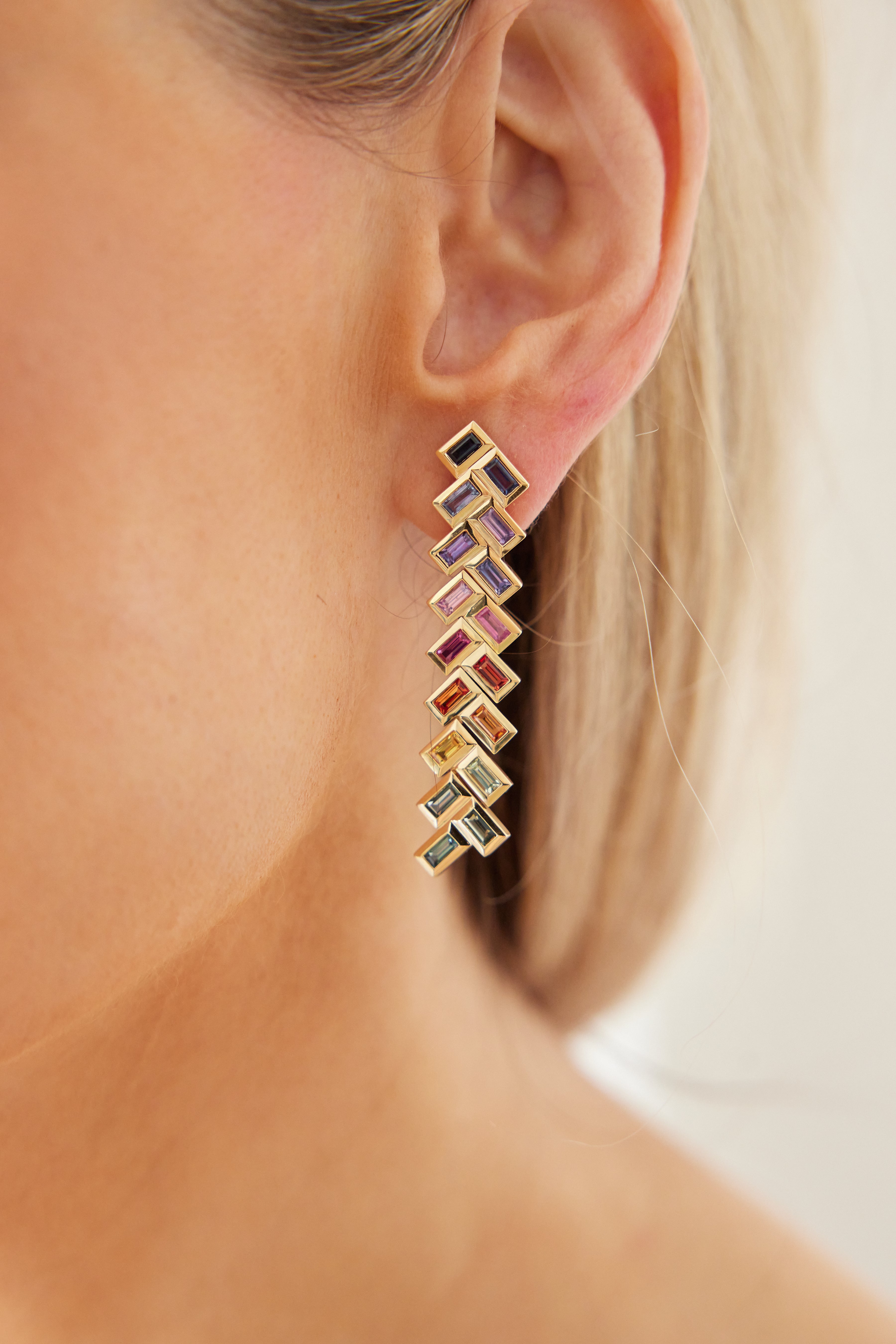 Close-up of a woman's ear wearing Chevron Drop Earrings in yellow gold, featuring a geometric zigzag design with multicolor baguette cut sapphires—purple, red, and green. A softly blurred background adds elegance.