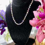The Chroma Kaleidoscope Mixed Shape Sapphire and Diamond Tennis Necklace in yellow gold is displayed on a black velvet stand, surrounded by vibrant pink and purple flowers in vases, highlighting its dazzling sapphire array.