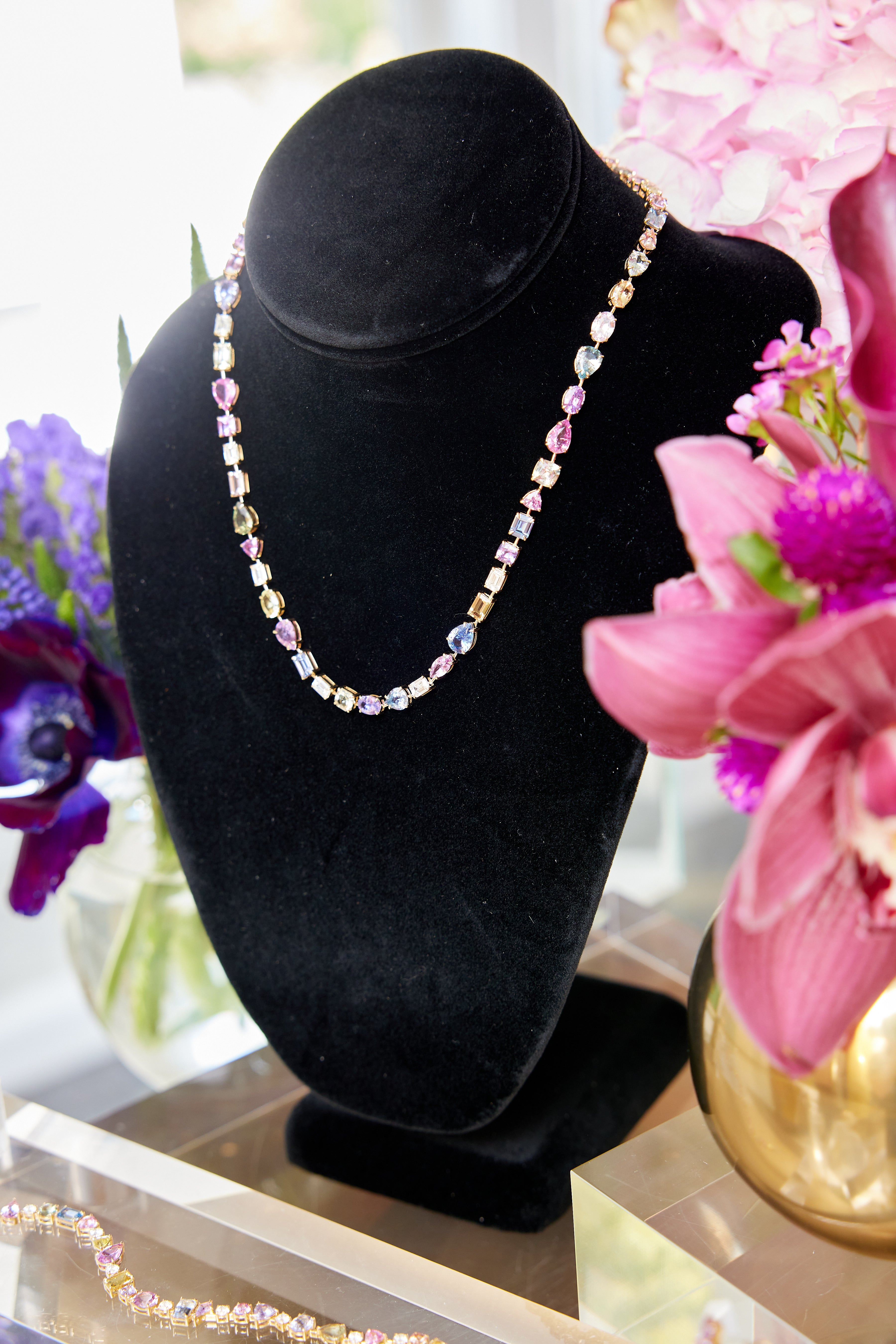 The Chroma Kaleidoscope Mixed Shape Sapphire and Diamond Tennis Necklace in yellow gold is displayed on a black velvet stand, surrounded by vibrant pink and purple flowers in vases, highlighting its dazzling sapphire array.