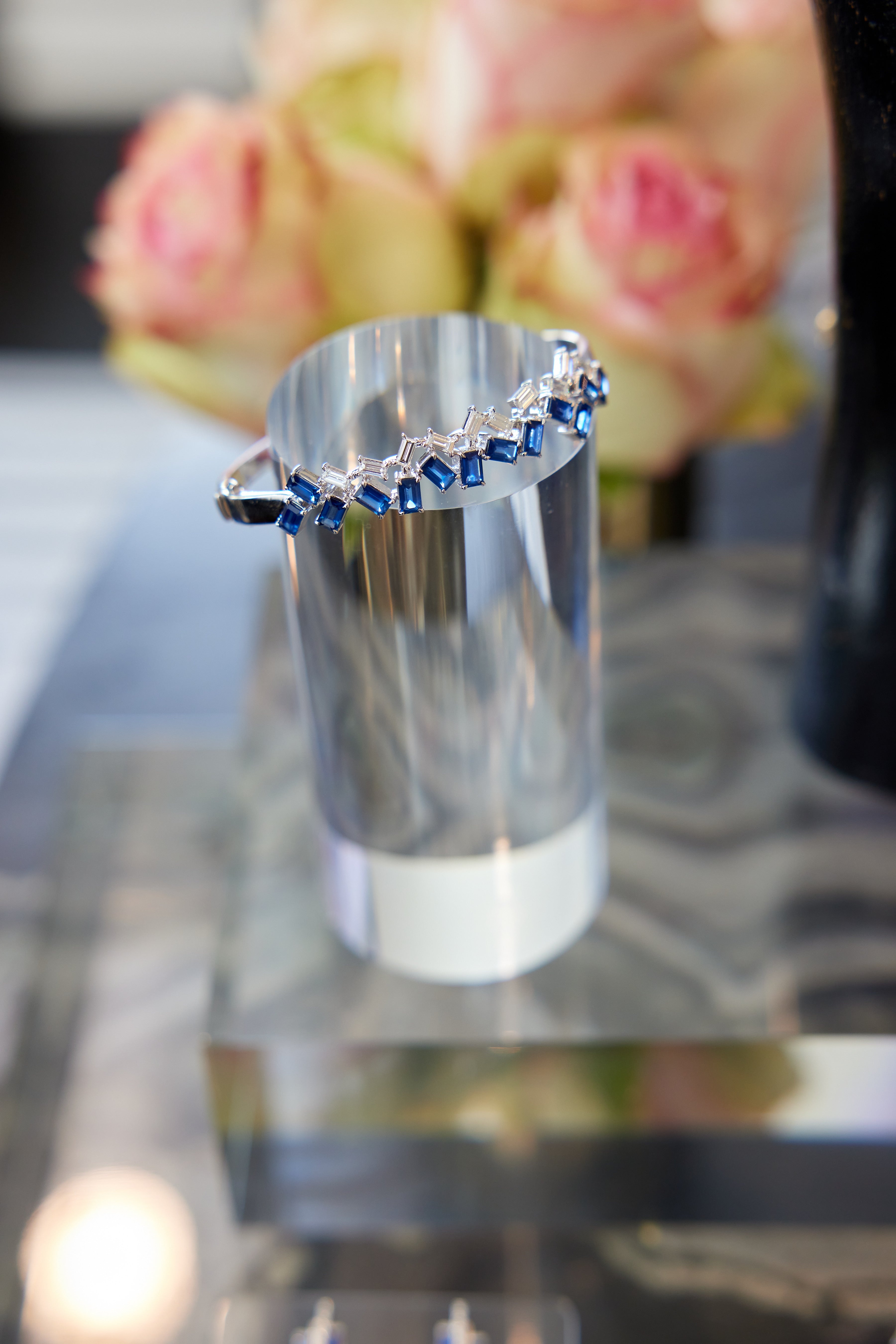 The Chroma Empire Alternating Emerald Cut Sapphire and Diamond Bangle in White Gold sparkles on a stand, with blurred pink and cream roses in the background enhancing the jewelry's luminous elegance.