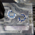 The Chroma Empire Alternating Sapphire and Diamond Front-to-Back Earrings feature crescent-shaped blue sapphires and diamonds in 18K white gold, shown on a clear acrylic stand with a blurred backdrop.