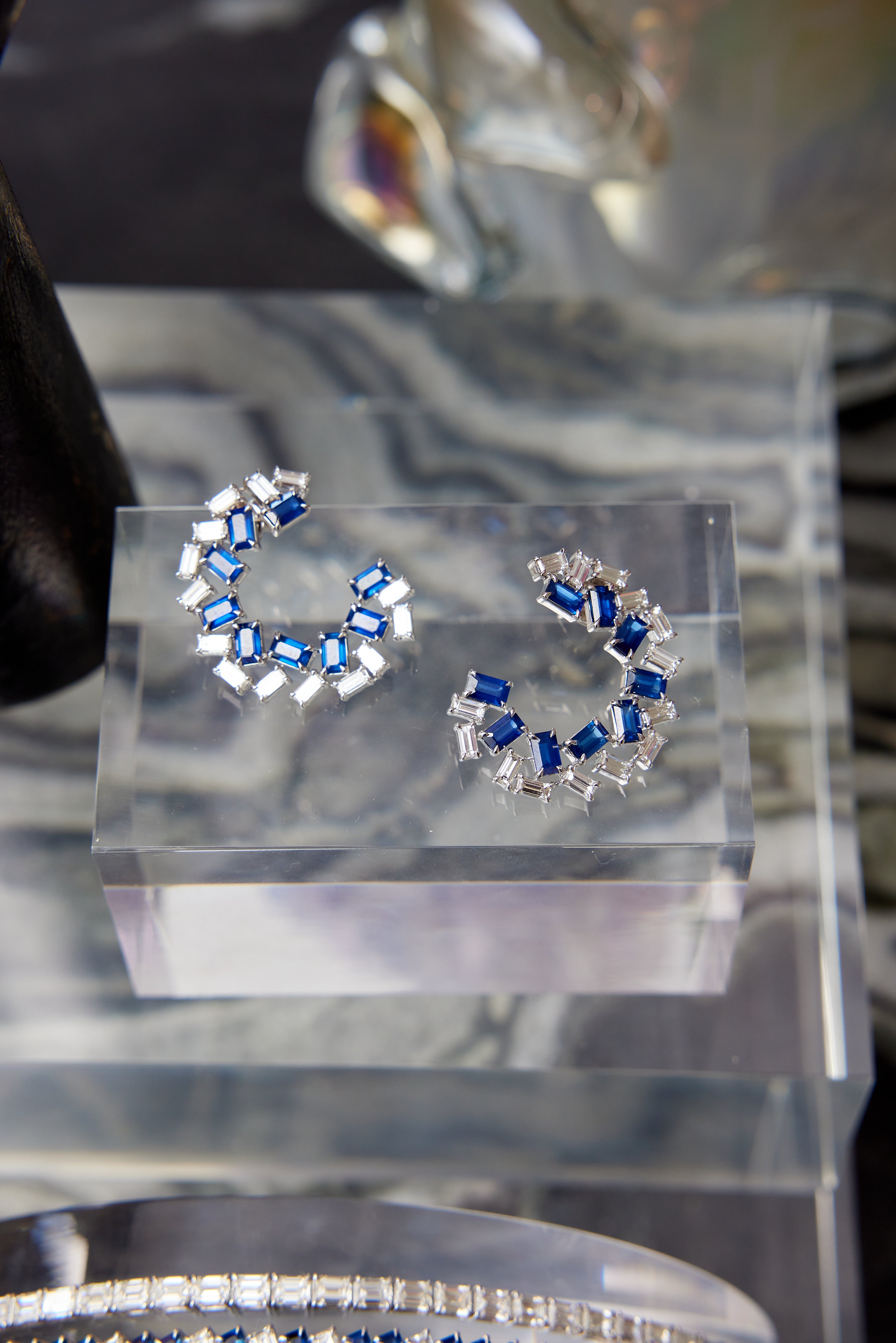 The Chroma Empire Alternating Sapphire and Diamond Front-to-Back Earrings feature crescent-shaped blue sapphires and diamonds in 18K white gold, shown on a clear acrylic stand with a blurred backdrop.