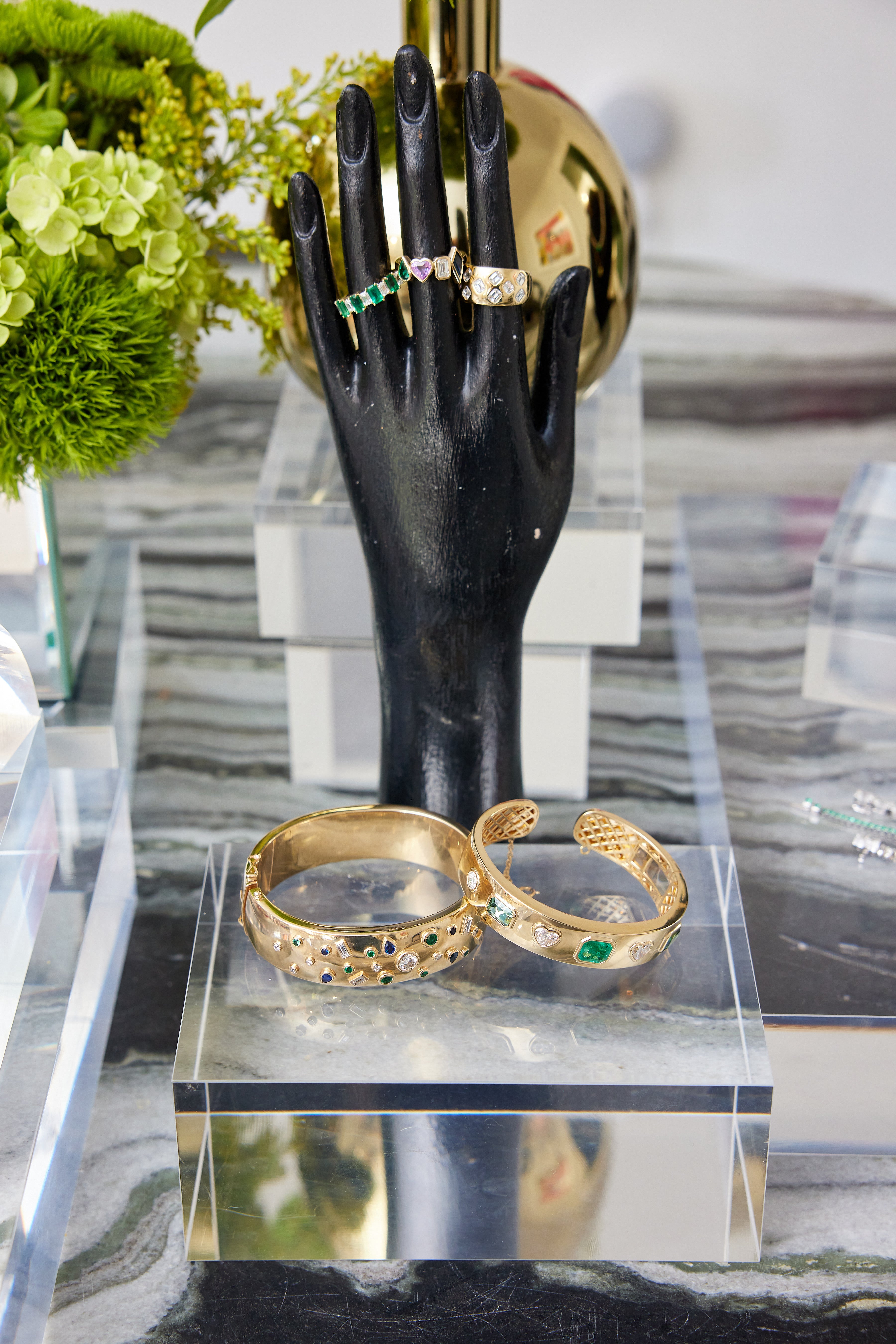 A black mannequin hand displays the Chroma Alternating Emerald and Diamond Half Eternity Band with gold rings. In front, two gold gem-accented bracelets rest on a clear acrylic stand atop striped marble, with green flowers in the background.