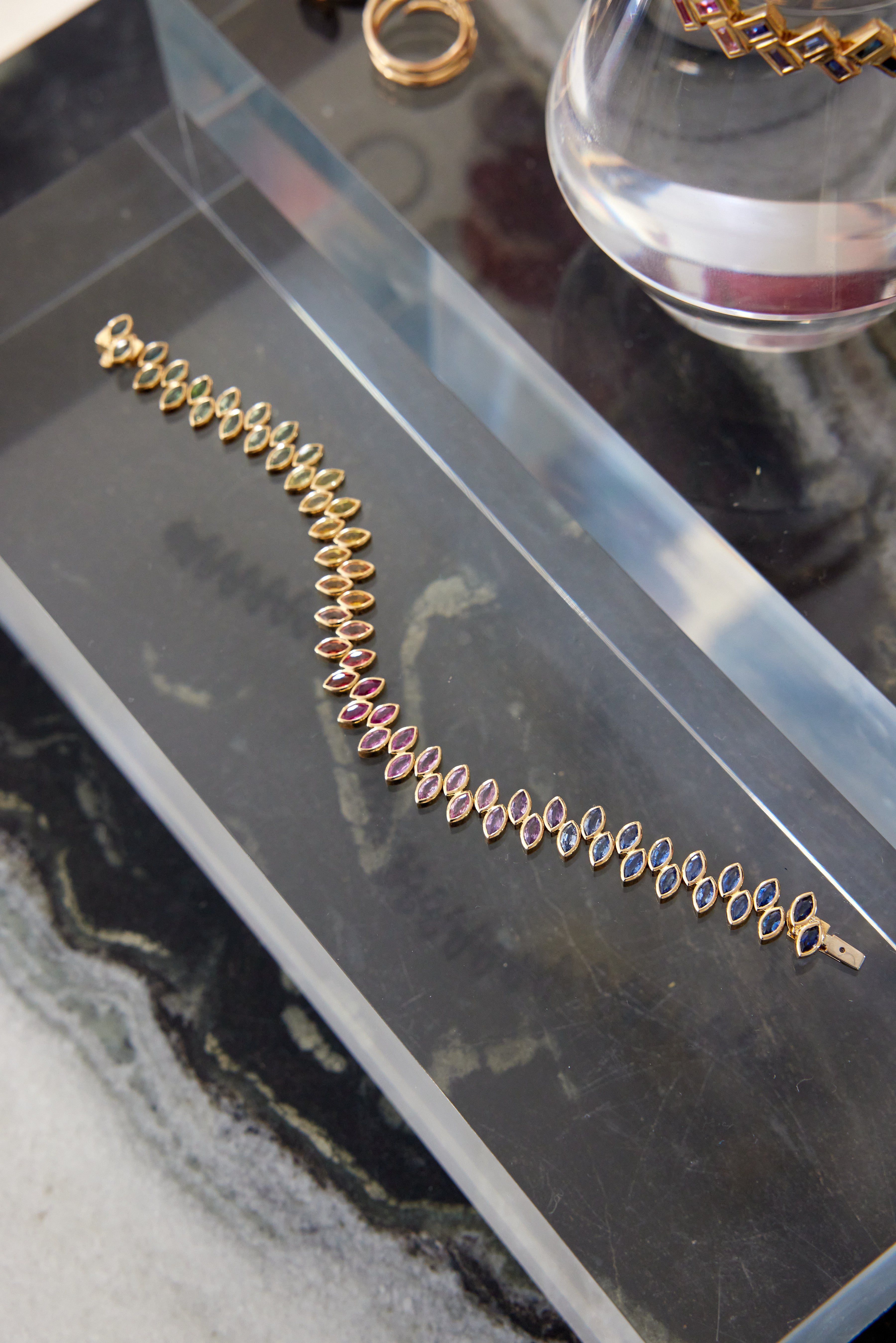 The Chroma Marching Alternating Bezel Set Marquise Sapphire Tennis Bracelet in yellow gold is displayed on a clear rectangular tray, with other jewelry pieces visible in the background.