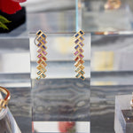 The Chevron Drop Earrings in Yellow Gold with multicolor baguette cut sapphires are displayed upright on a clear acrylic stand, with other jewelry and a red flower softly blurred in the background.