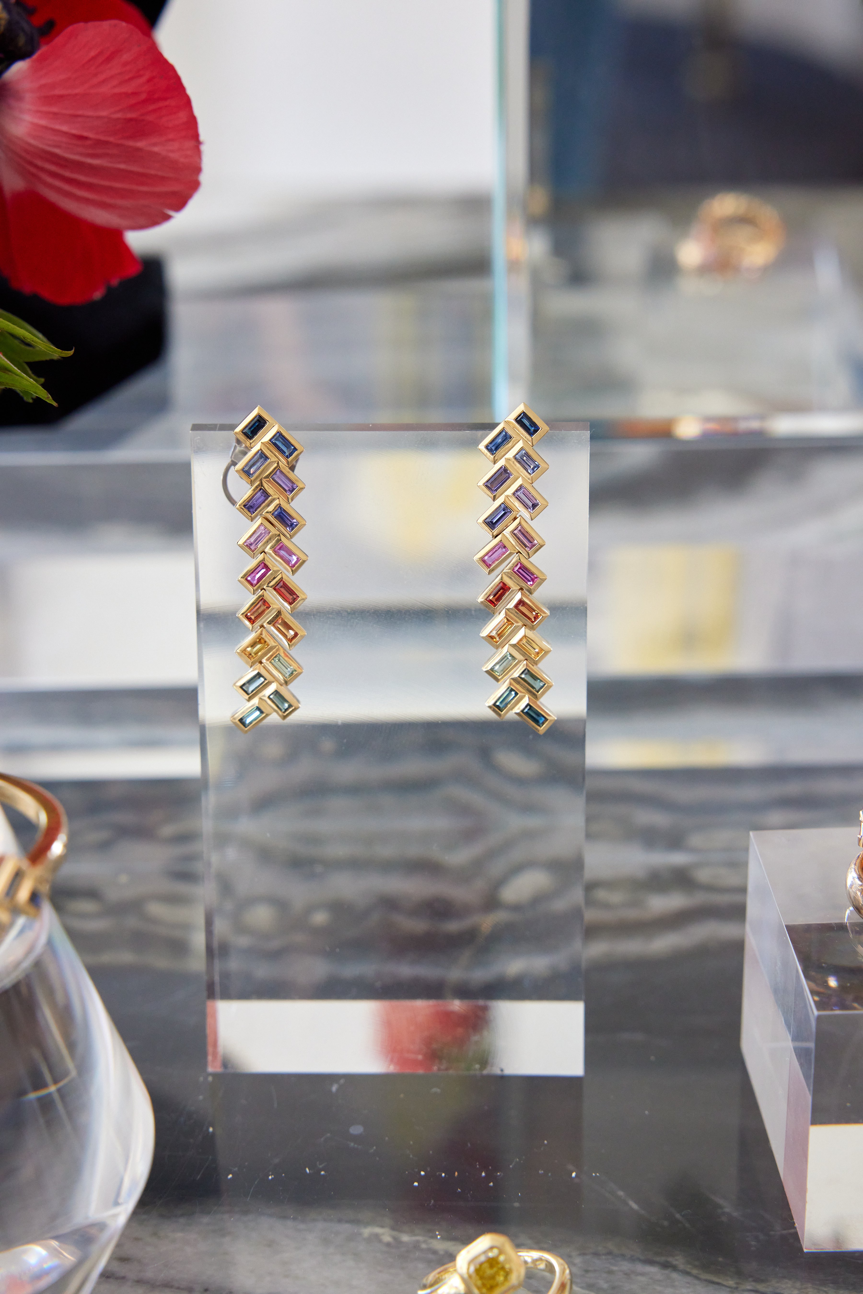 The Chevron Drop Earrings in Yellow Gold with multicolor baguette cut sapphires are displayed upright on a clear acrylic stand, with other jewelry and a red flower softly blurred in the background.