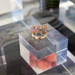 The Chroma Chevron Bezel Set Ring, featuring a colorful woven pattern of multicolor baguette sapphires in gold, is displayed on a clear acrylic cube with other jewelry and blurred background elements visible around it.