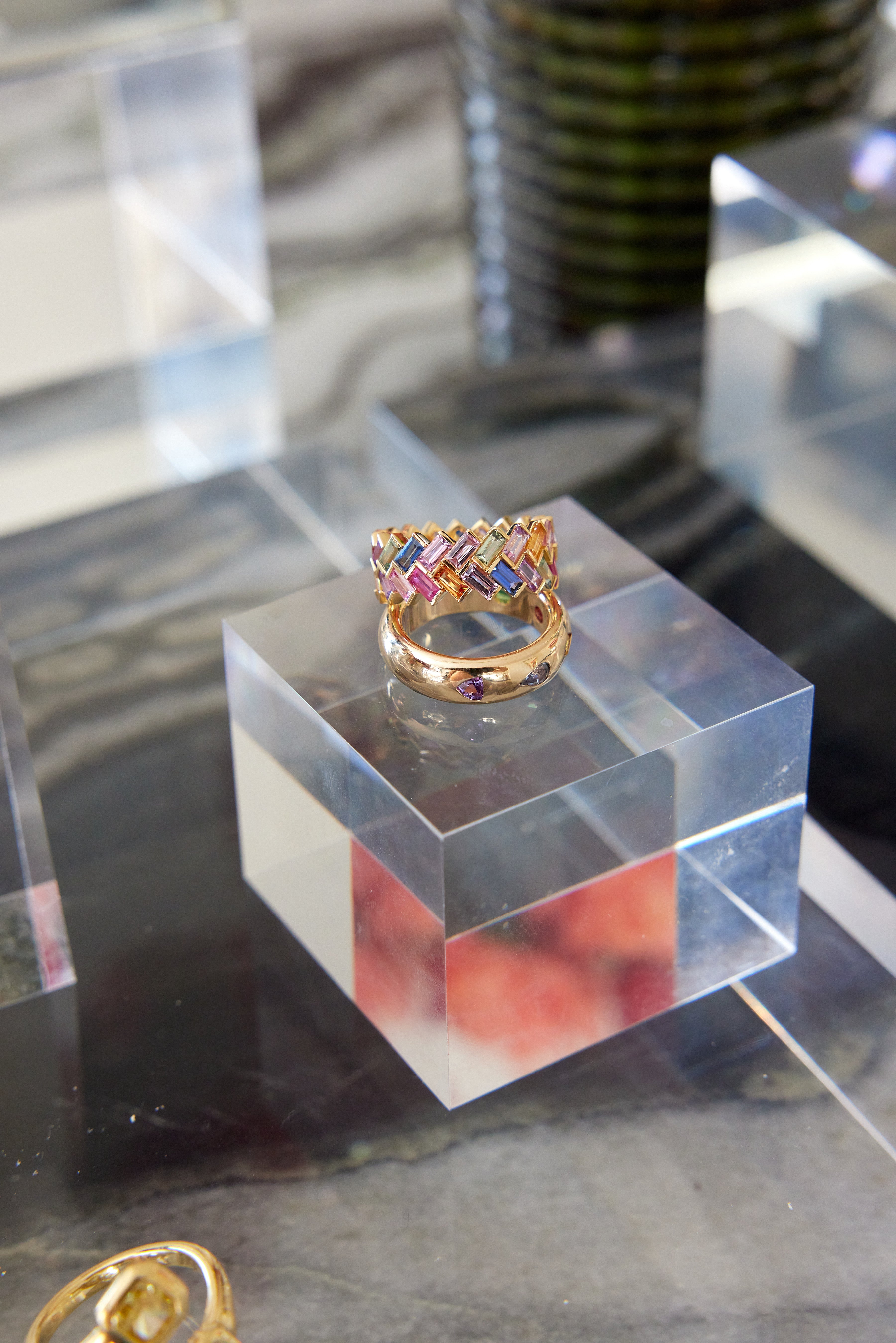 The Chroma Chevron Bezel Set Ring, featuring a colorful woven pattern of multicolor baguette sapphires in gold, is displayed on a clear acrylic cube with other jewelry and blurred background elements visible around it.